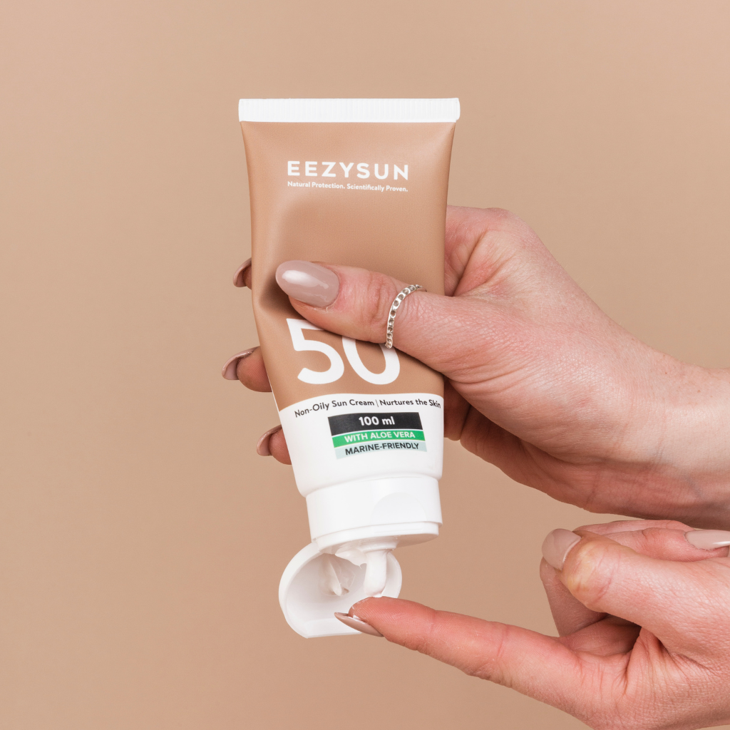 Image of the EEZYSUN SPF 50 in a tube product on a peach background with a hand squeezing the cream out onto their finger.